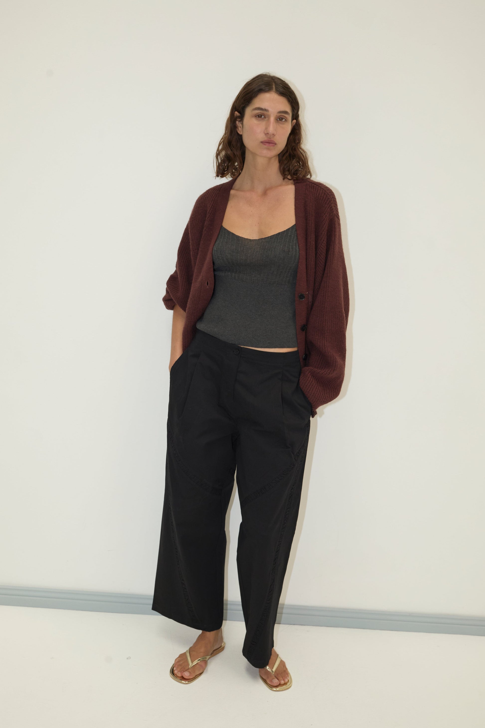 A woman with wavy brown hair stands against a plain light wall, wearing a Flounce Cardigan - Burgundy by Deiji Studios over a gray tank, loose black pants, and gold sandals, her hands in her pockets.