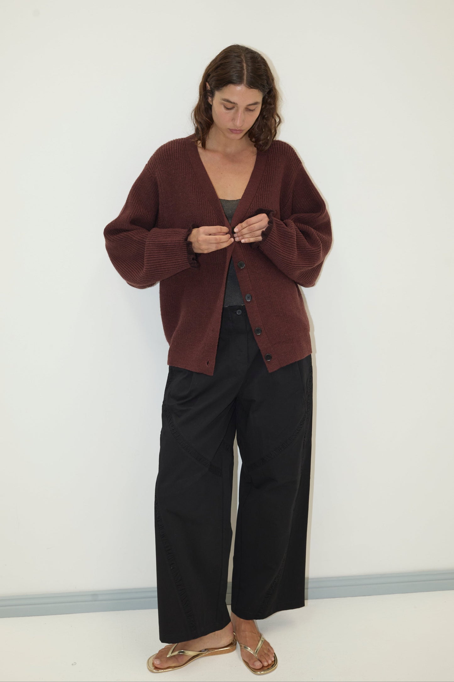 A woman stands against a white wall, wearing the Flounce Cardigan - Burgundy made from recycled acrylic over a gray top, black wide-leg pants, and gold sandals. She looks down as she fastens a button on her cardigan.