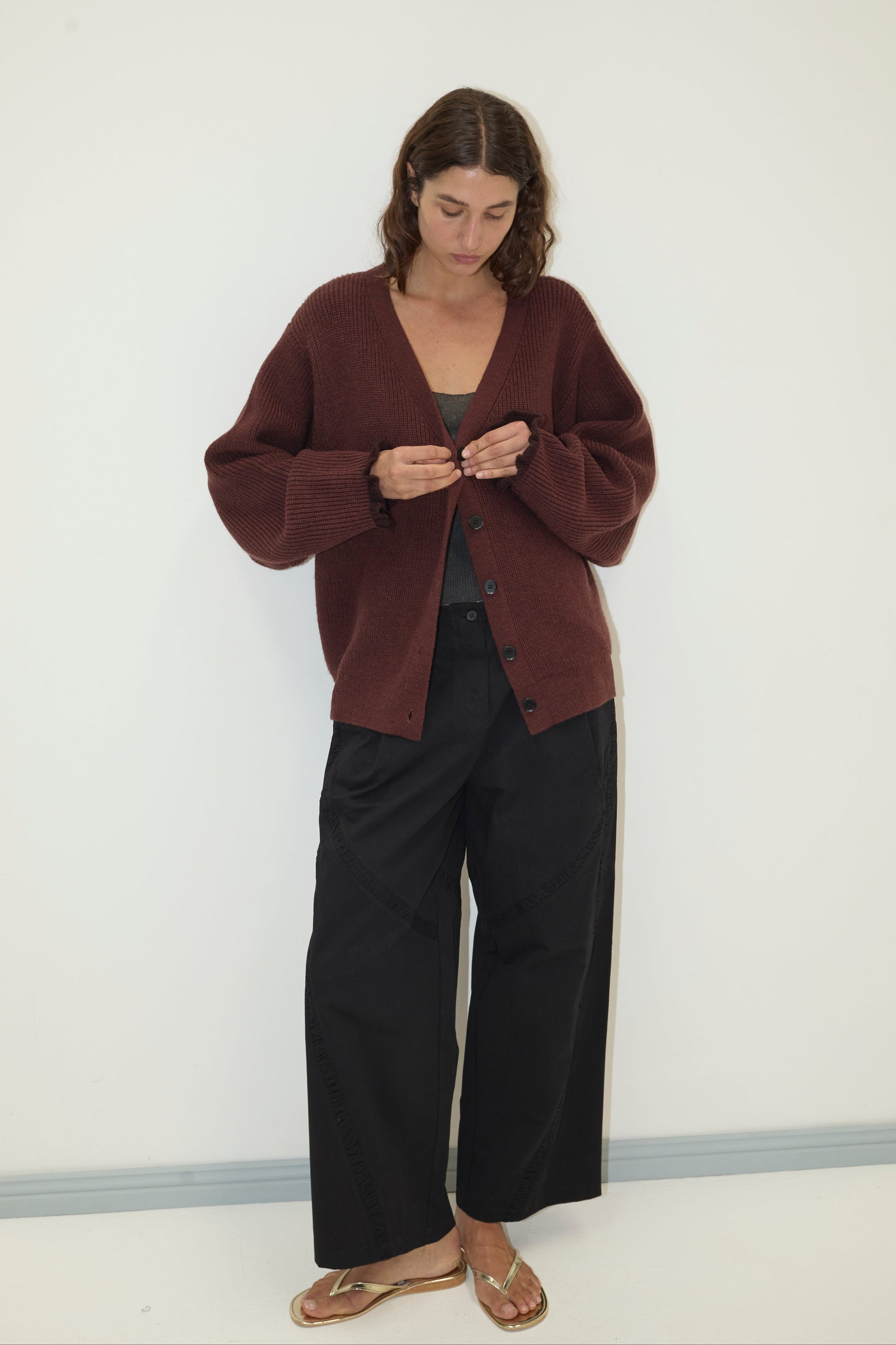 A woman stands against a white wall, wearing the Flounce Cardigan - Burgundy made from recycled acrylic over a gray top, black wide-leg pants, and gold sandals. She looks down as she fastens a button on her cardigan.