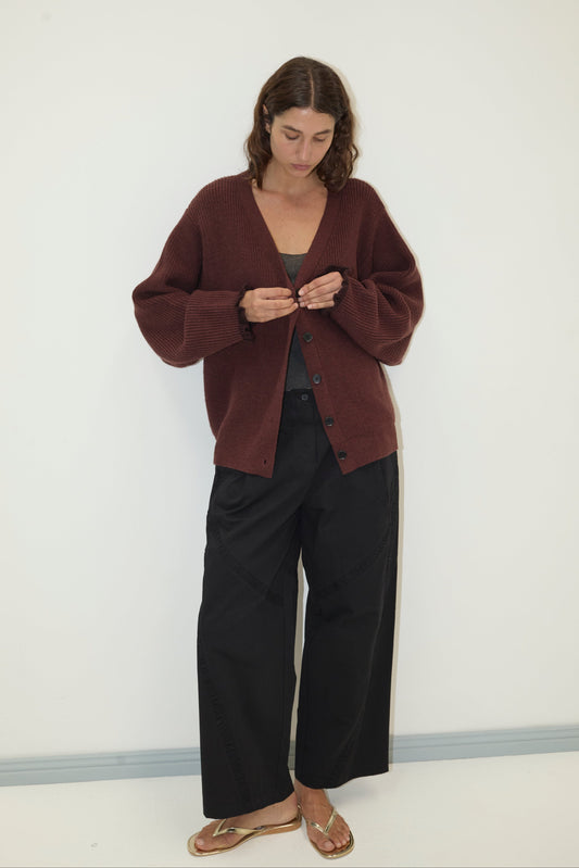 A woman stands against a white wall, wearing the Flounce Cardigan - Burgundy made from recycled acrylic over a gray top, black wide-leg pants, and gold sandals. She looks down as she fastens a button on her cardigan.