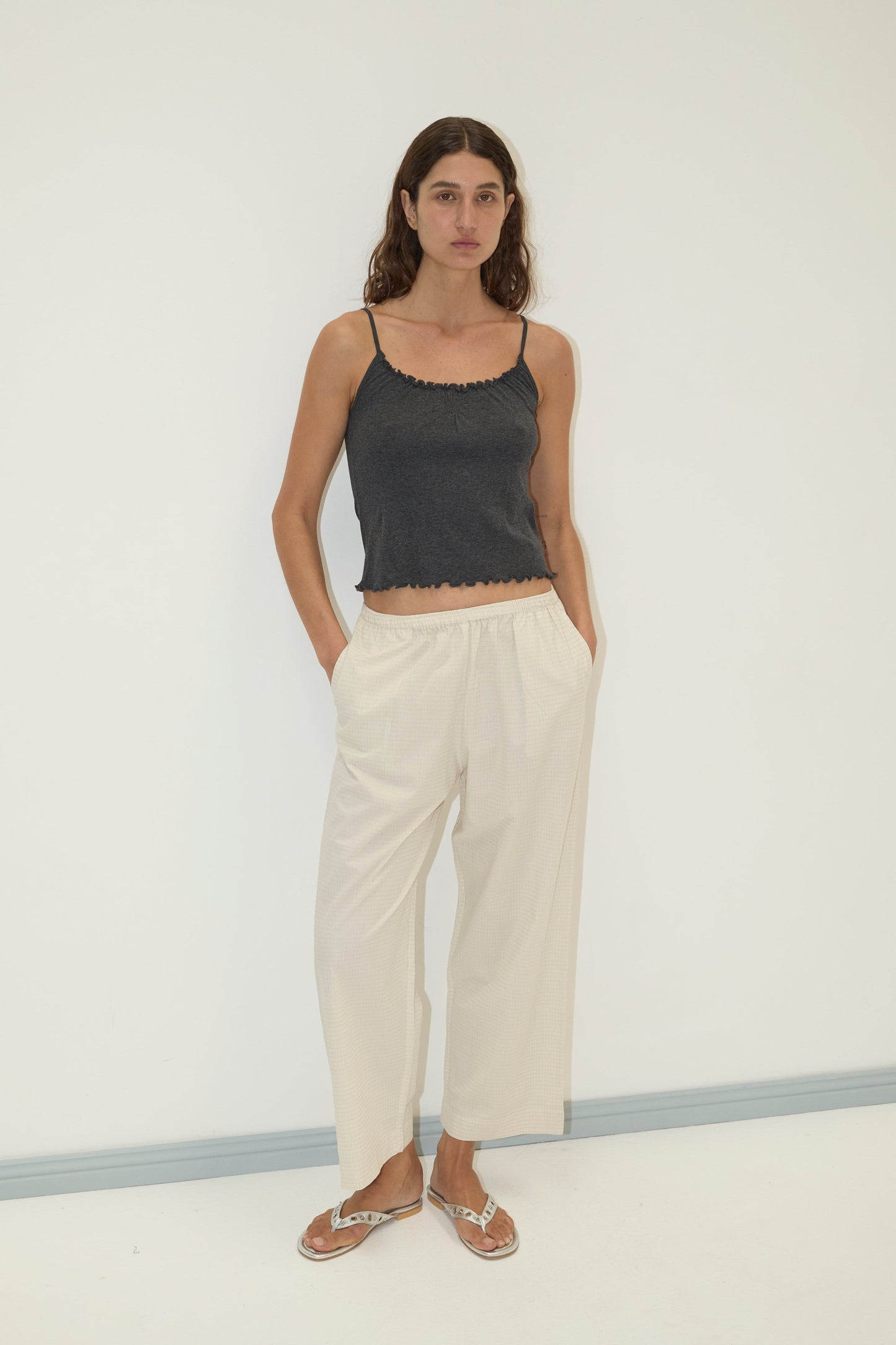 A woman with long, wavy hair stands against a plain white wall wearing the Flounce Tank - Grey by Deiji Studios, loose light-colored pants, and flip-flops, with her hands in her pockets.