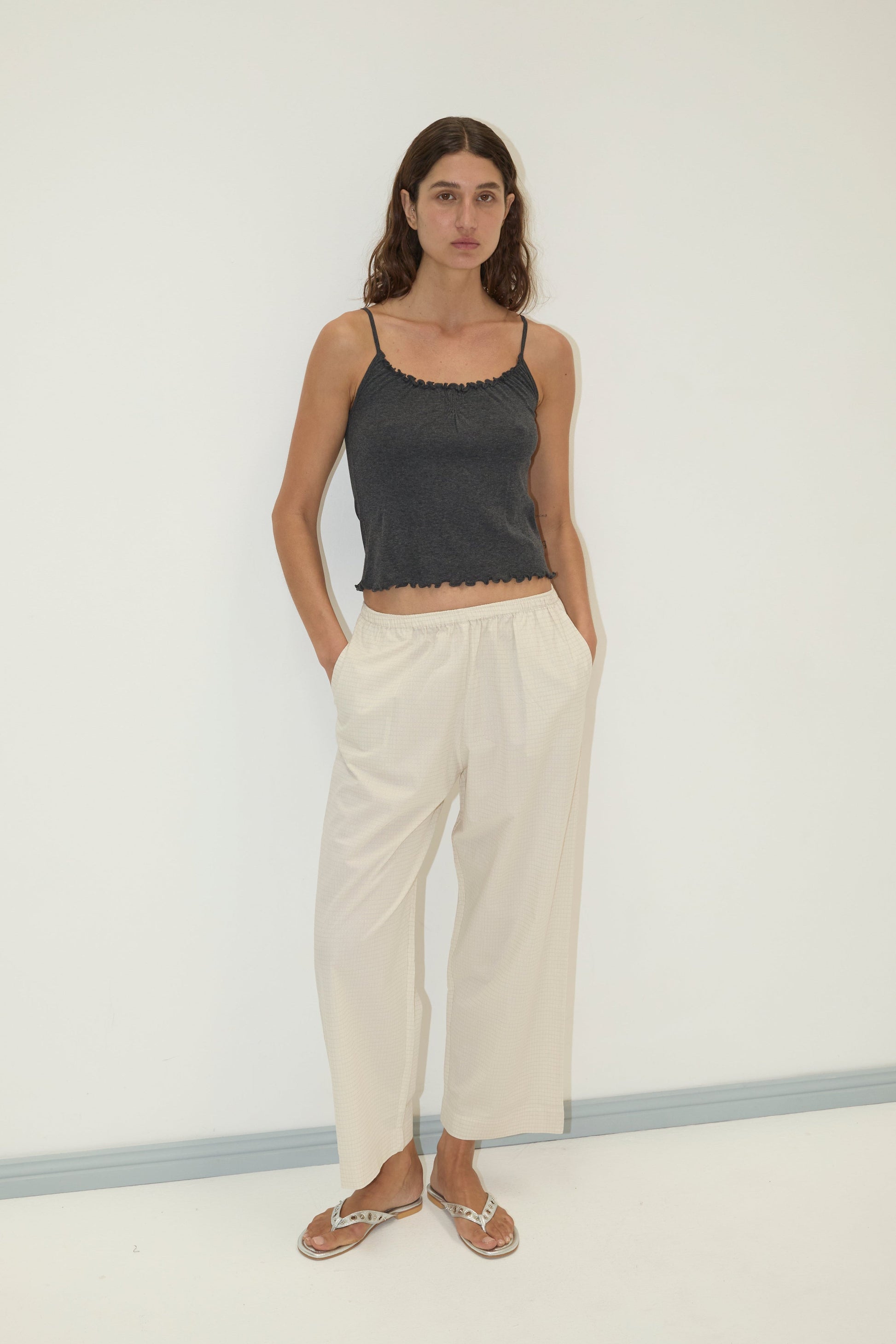 A woman with long, wavy hair stands against a plain white wall wearing the Flounce Tank - Grey by Deiji Studios, loose light-colored pants, and flip-flops, with her hands in her pockets.