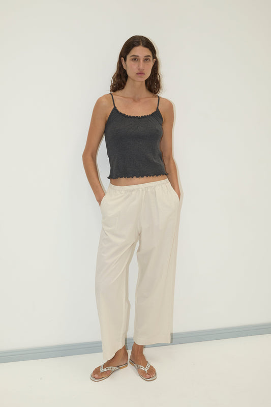 A woman with long, wavy hair stands against a plain white wall wearing the Flounce Tank - Grey by Deiji Studios, loose light-colored pants, and flip-flops, with her hands in her pockets.