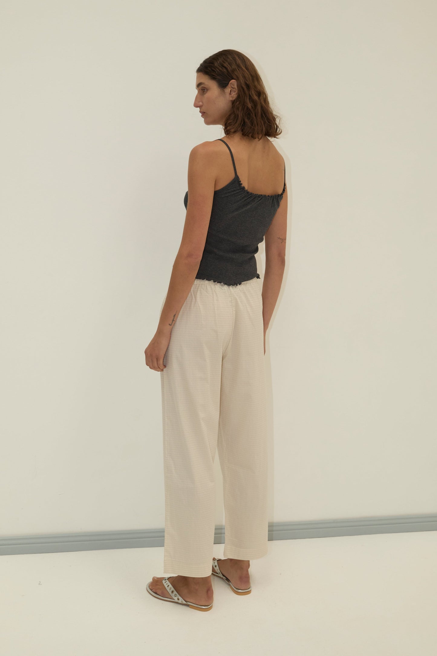 A woman with brown hair, wearing the Deiji Studios Flounce Tank in grey, loose light beige pants, and sandals stands facing slightly away in a minimalist setting with a light background.