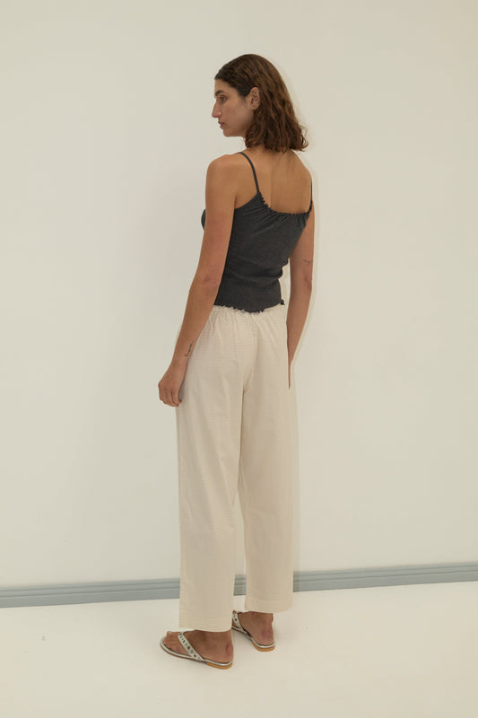 A woman with brown hair, wearing the Deiji Studios Flounce Tank in grey, loose light beige pants, and sandals stands facing slightly away in a minimalist setting with a light background.