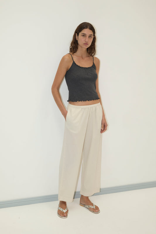 A woman with long brown hair stands against a white wall, wearing the Ease Trouser in cream, a dark gray spaghetti strap top, and sandals. With a neutral expression and one hand in her pocket, she exudes effortless style.