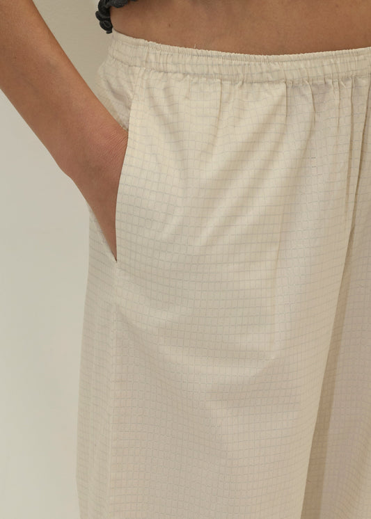 Someone wears the Deiji Studios Ease Trouser in Bone Check, featuring a loose fit and subtle cream checkered pattern, with a hand in the pocket against a plain light background.