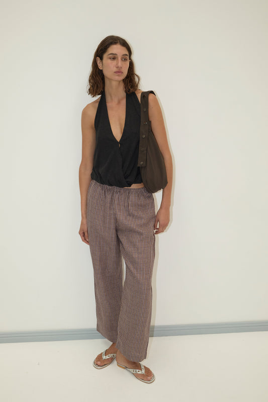 A woman with wavy brown hair and a neutral expression stands against a plain light-colored wall wearing Deiji Studios' linen ease trouser in russet check, a sleeveless deep V-neck black top, silver sandals, and a brown shoulder bag.