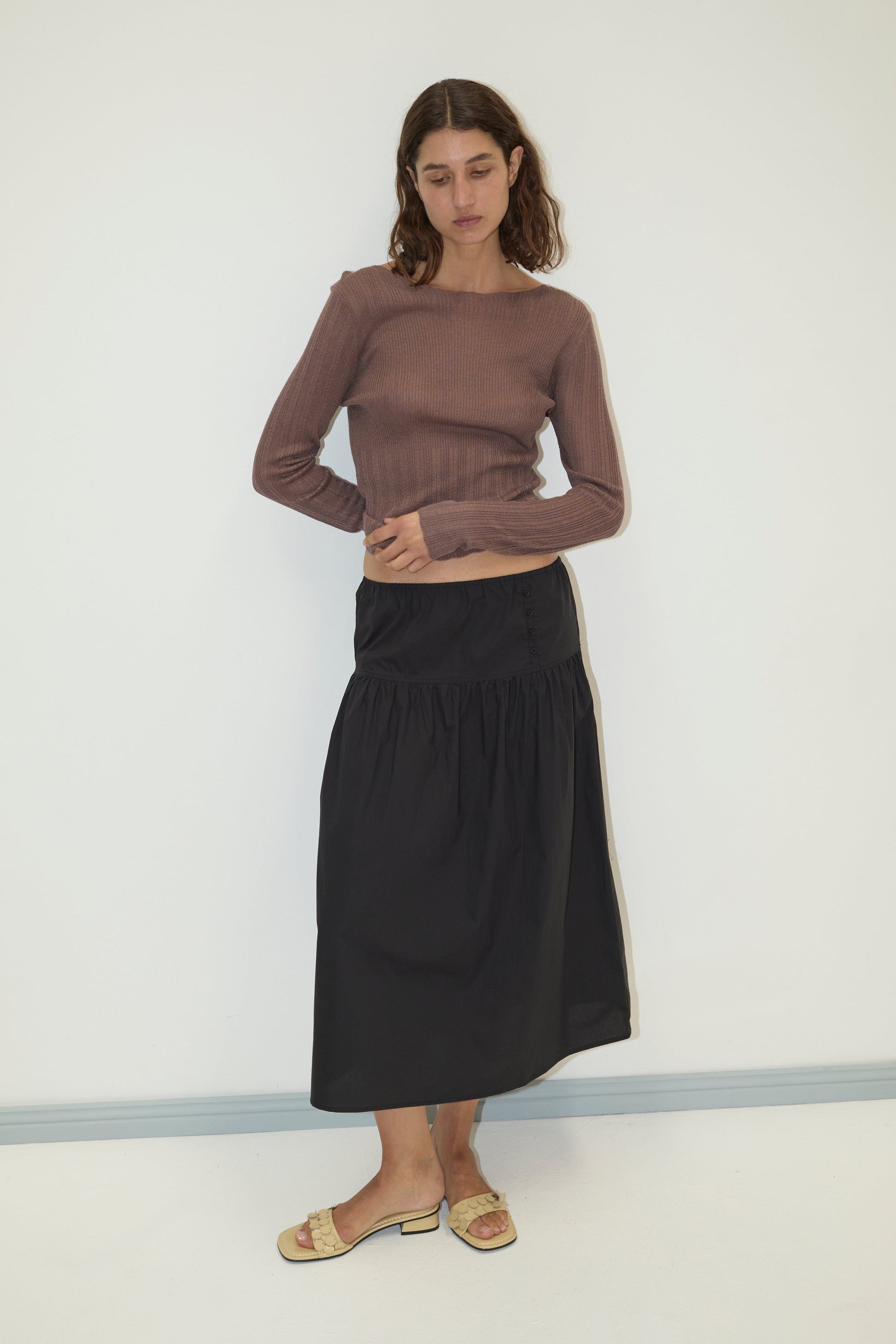 A woman with wavy brown hair wears a long-sleeved ribbed brown top, the Deiji Studios Button Skirt in black, and beige open-toe slide sandals, standing against a plain white wall.