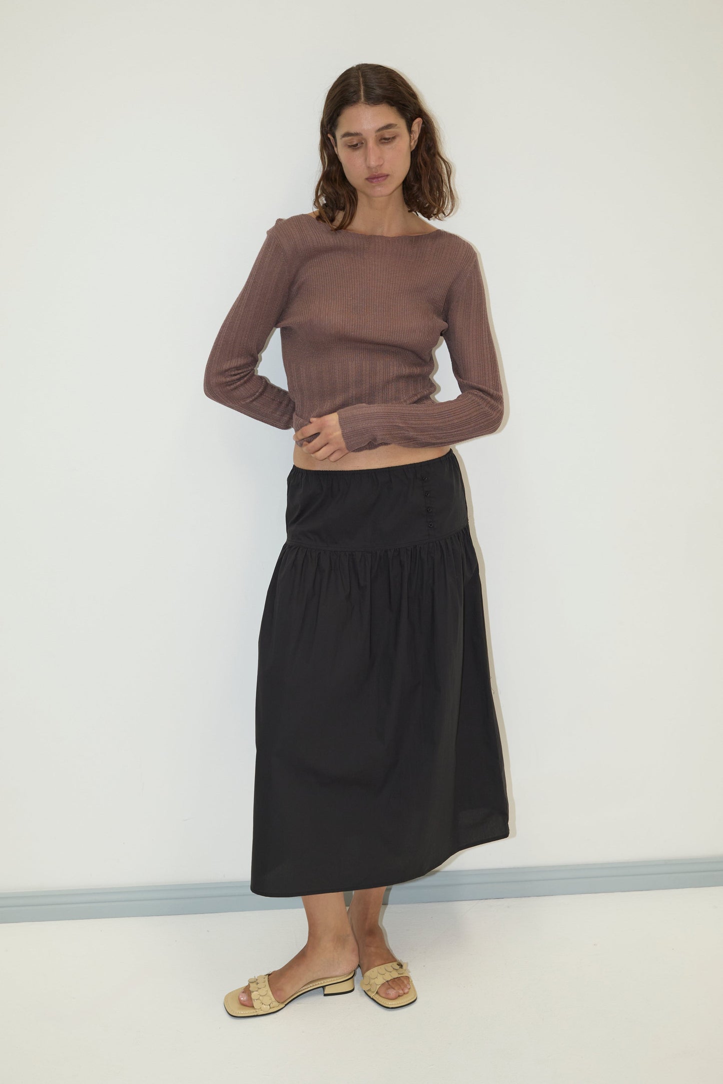 A woman wearing a fitted mauve ribbed long-sleeve top, a black Button Skirt, and beige strappy sandals stands against a white wall.
