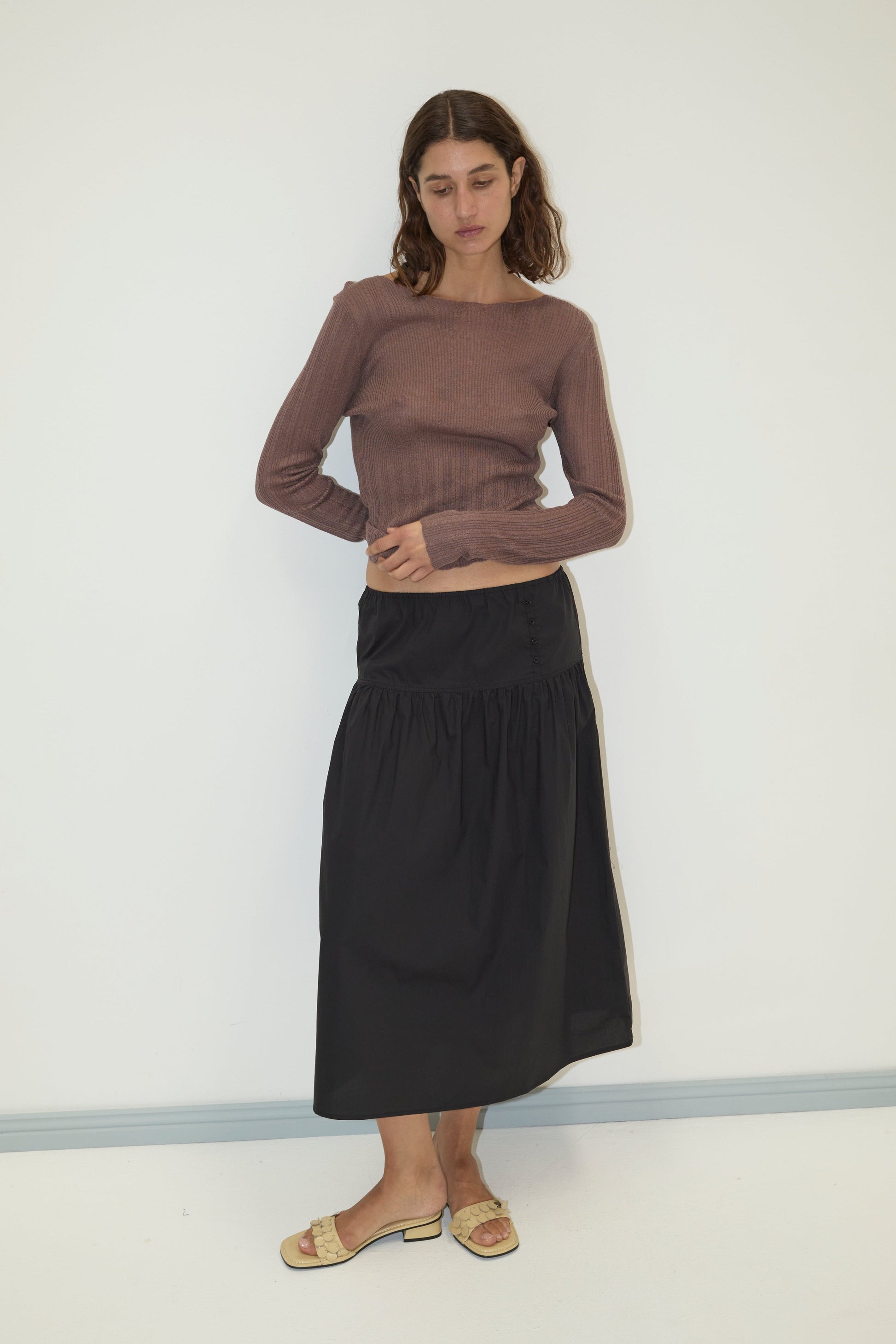 A woman with shoulder-length brown hair and a neutral expression wears the Deiji Studios Open Work Longsleeve in Hazel, paired with a black midi skirt and sandals against a plain white wall, reflecting minimalist style.