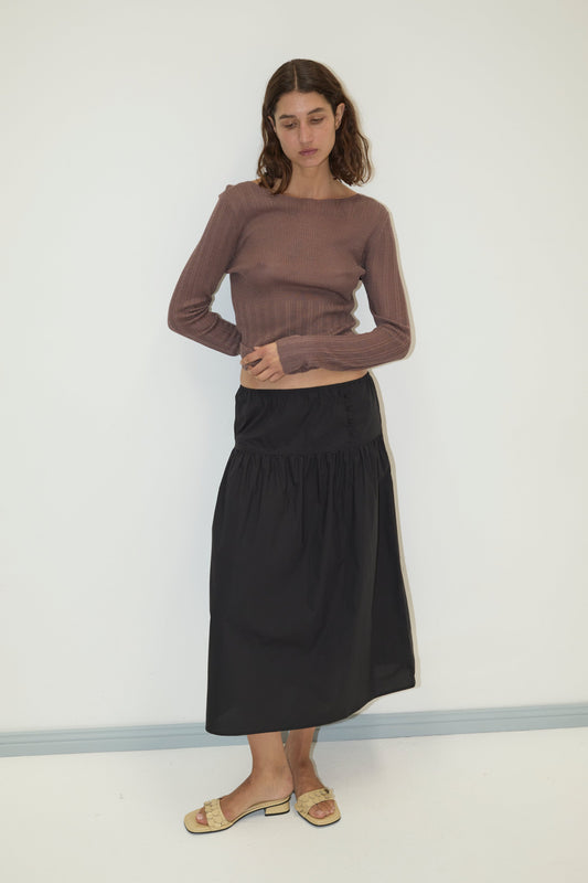 A woman with shoulder-length brown hair and a neutral expression wears the Deiji Studios Open Work Longsleeve in Hazel, paired with a black midi skirt and sandals against a plain white wall, reflecting minimalist style.
