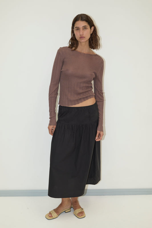 A woman with wavy brown hair and a neutral expression stands against a plain white wall, wearing the Open Work Longsleeve - Hazel by Deiji Studios, a black midi skirt, and beige slide sandals.