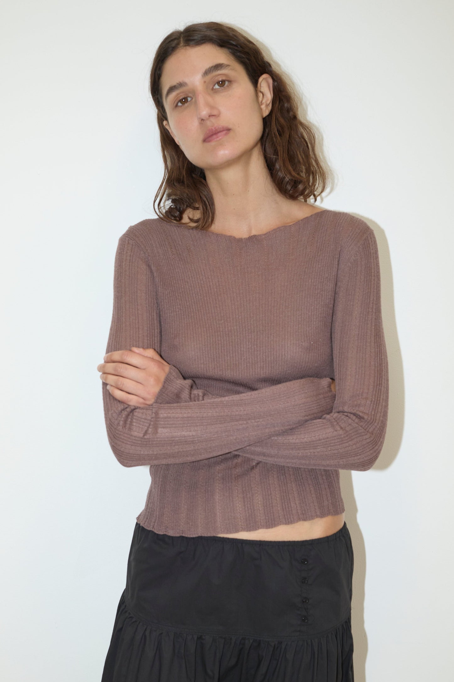 A woman with wavy brown hair stands against a plain white wall, wearing the Open Work Longsleeve - Hazel and a black skirt, her arms crossed and her expression neutral.