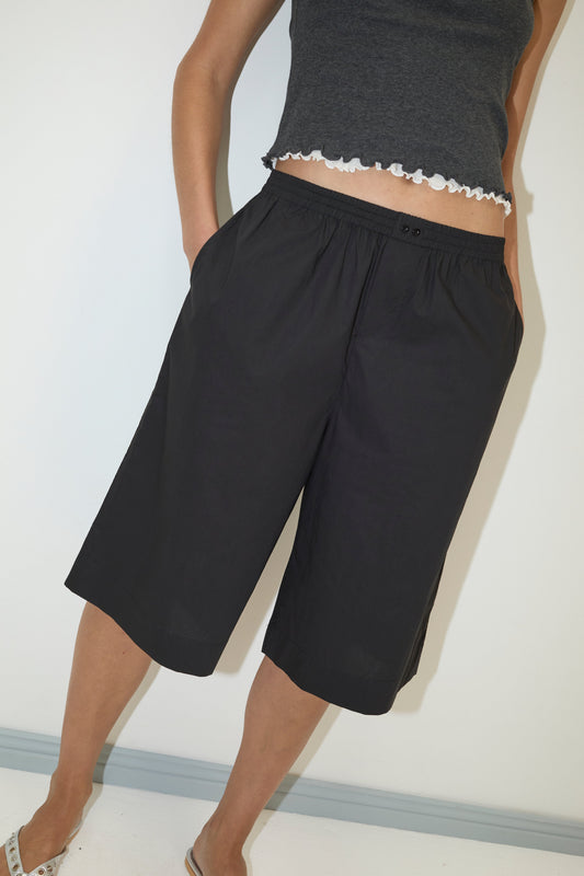 Wearing the Long Lounger - Black by Deiji Studios, a person with hands in pockets stands against a white wall, styled in a dark grey sleeveless top with ruffled hem and relaxed fit black knee-length shorts paired with sandals.