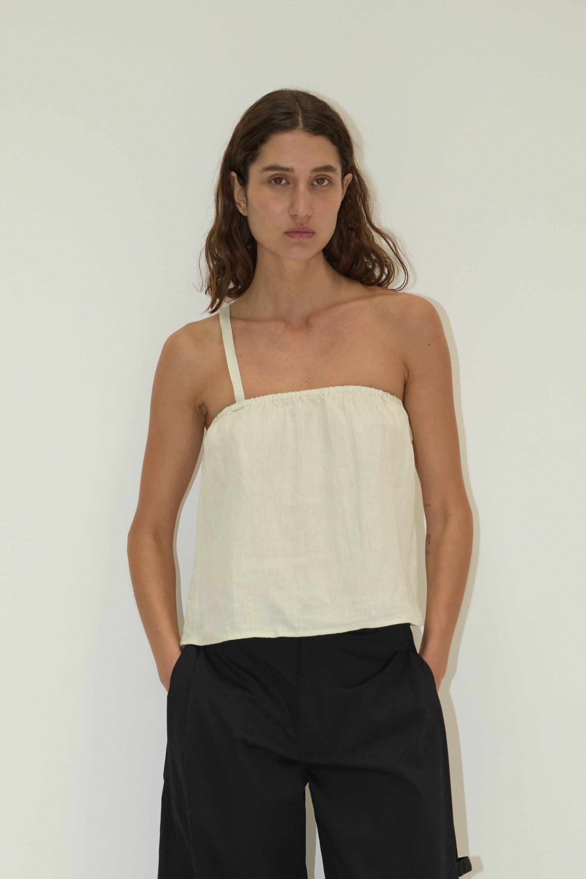 A woman with long brown hair wears a pastel pistachio Button Strap Top by Deiji Studios in organic French linen and loose black pants, standing against a plain white background with her hands in her pockets.