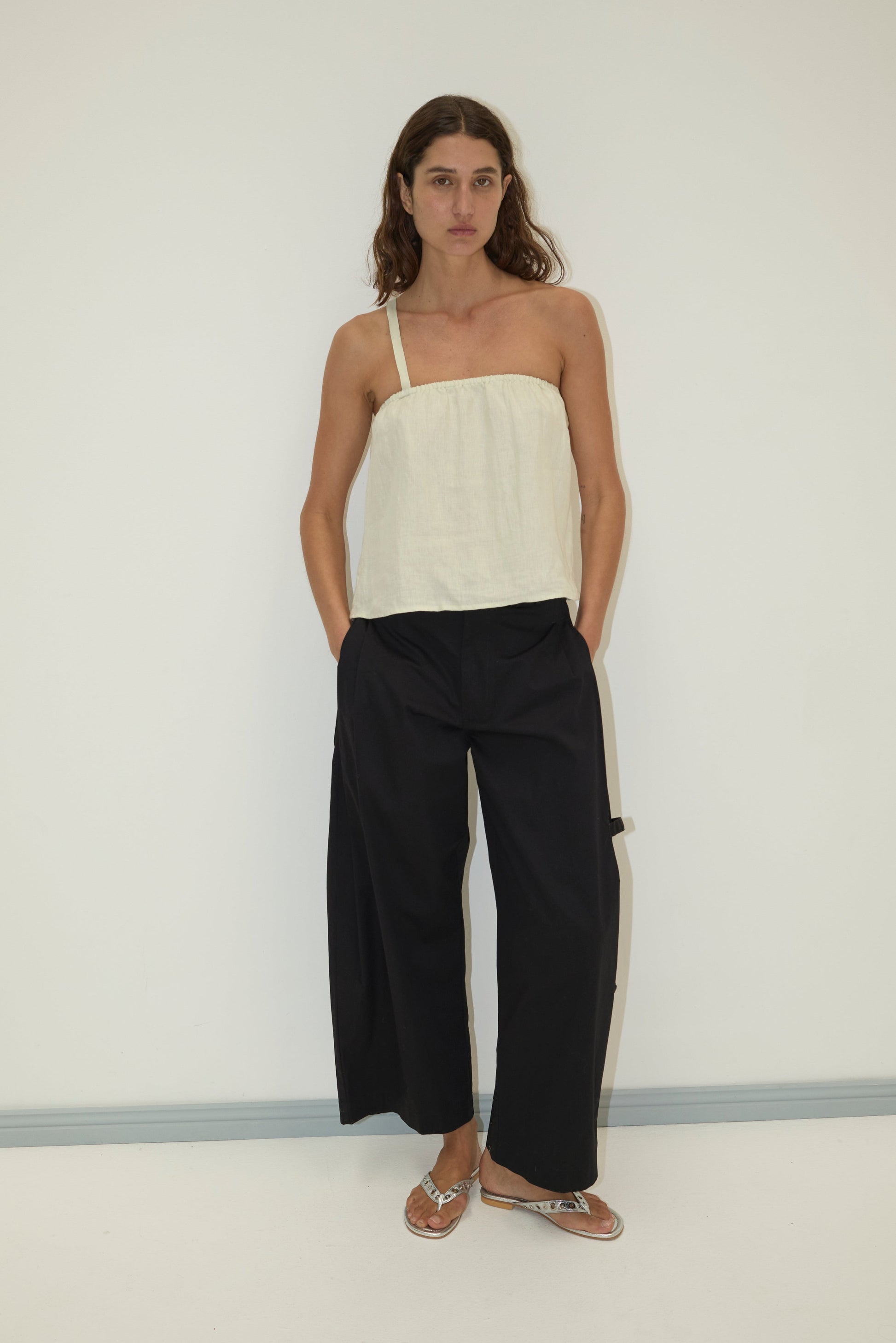 A woman stands against a white wall wearing a cream one-shoulder top, Deiji Studios’ Tab Pant in black wide-leg style, and silver flat sandals.