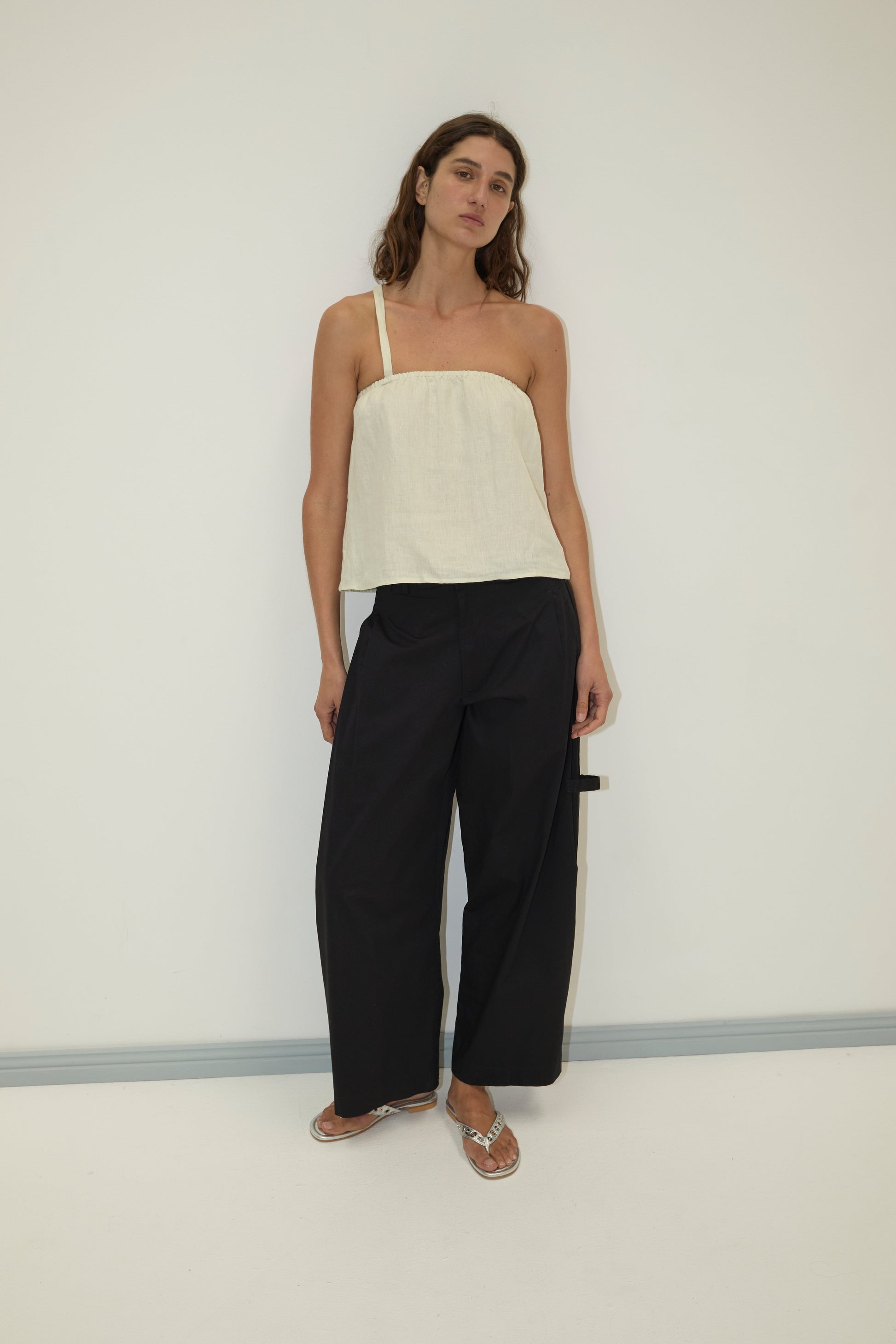 A woman stands against a plain wall wearing the Button Strap Top - Pastel Pistachio from Deiji Studios, crafted from organic French linen. She pairs it with loose black wide-leg pants and silver sandals, standing relaxed with one arm slightly bent.