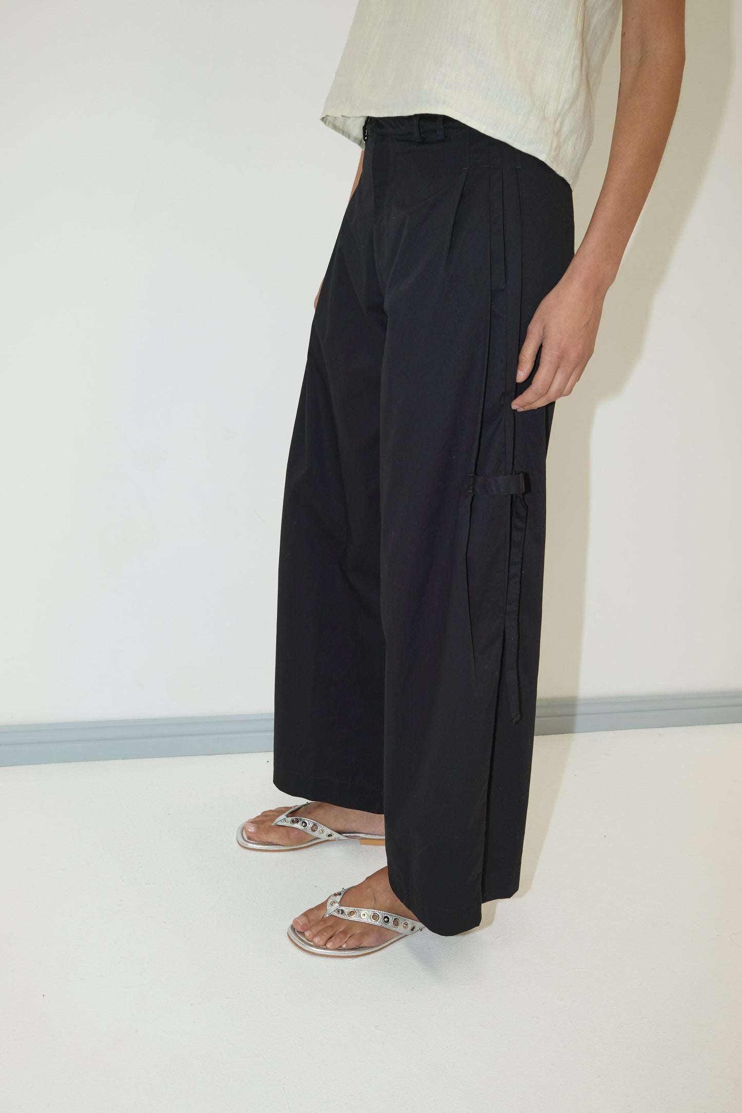 A person wearing the Tab Pant - Black from Deiji Studios, a light short-sleeve top, and silver sandals stands against a plain white background, with only the lower half of their body visible.