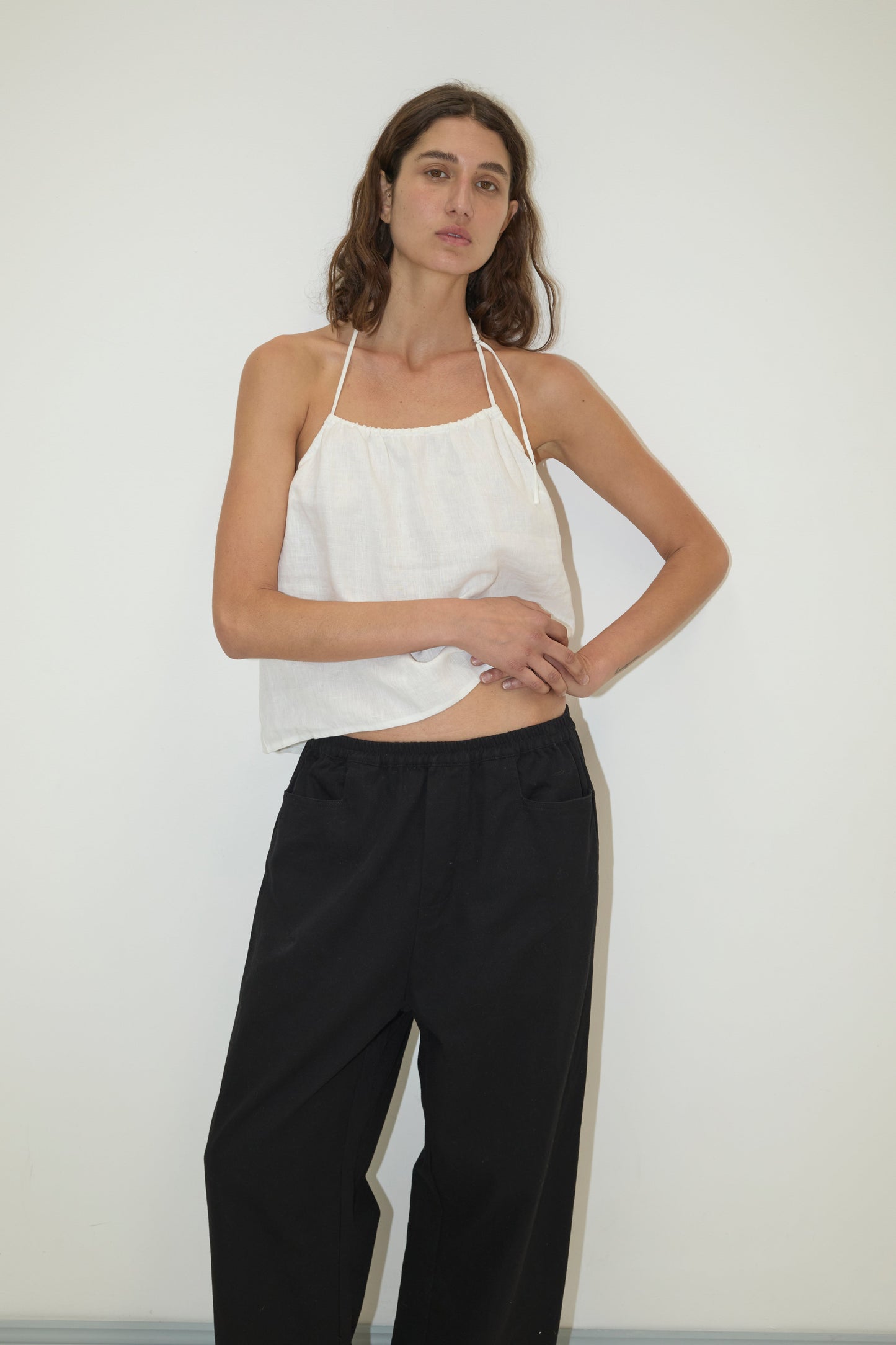 A woman with long brown hair wears a white halter top and the Square Pocket Pant in black, made from relaxed-fit organic cotton twill, standing against a plain white wall with one arm across her torso and the other on her hip.