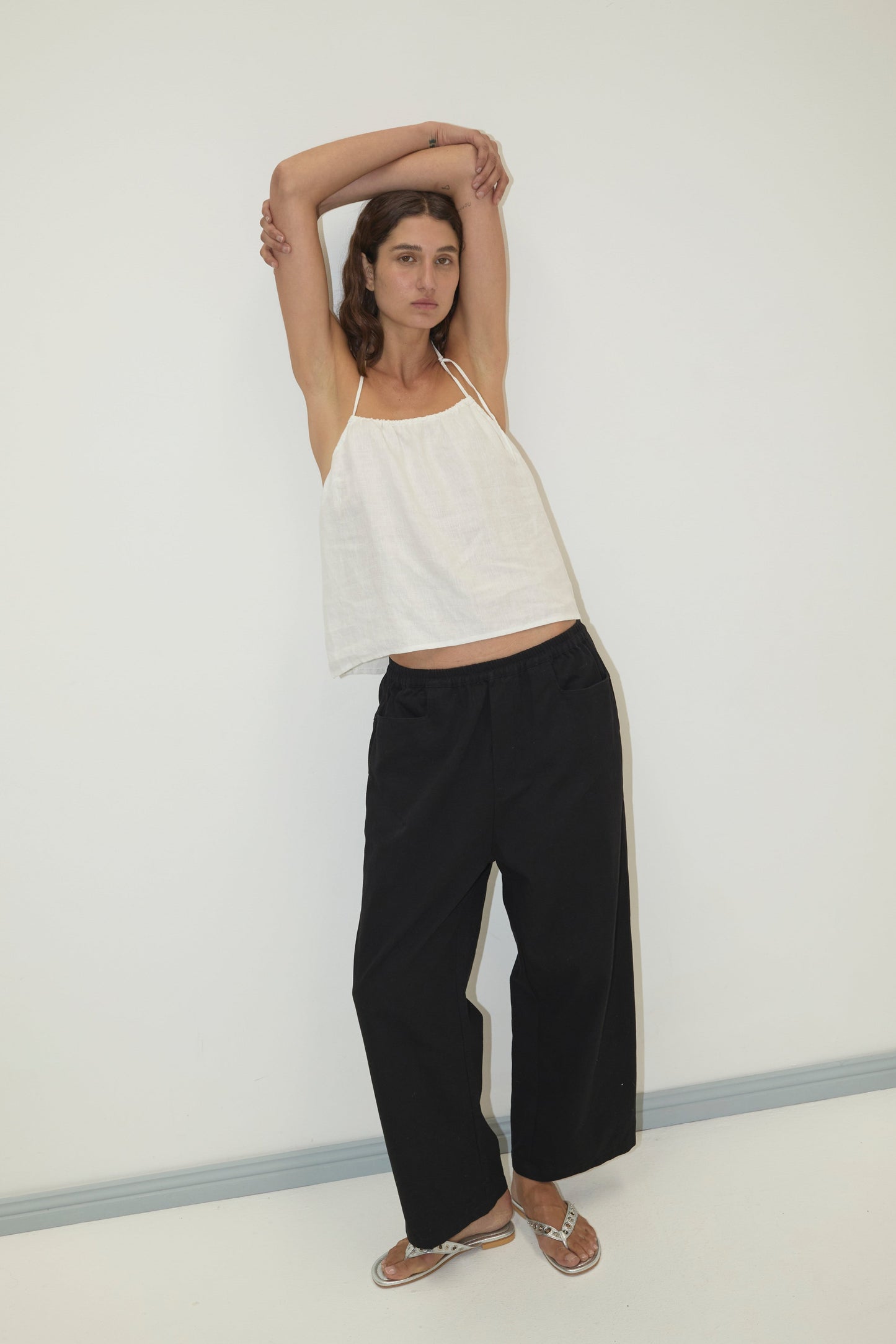 A person with long hair poses against a white wall, arms raised, wearing a loose white halter top, silver sandals, and the Square Pocket Pant in black organic cotton twill.