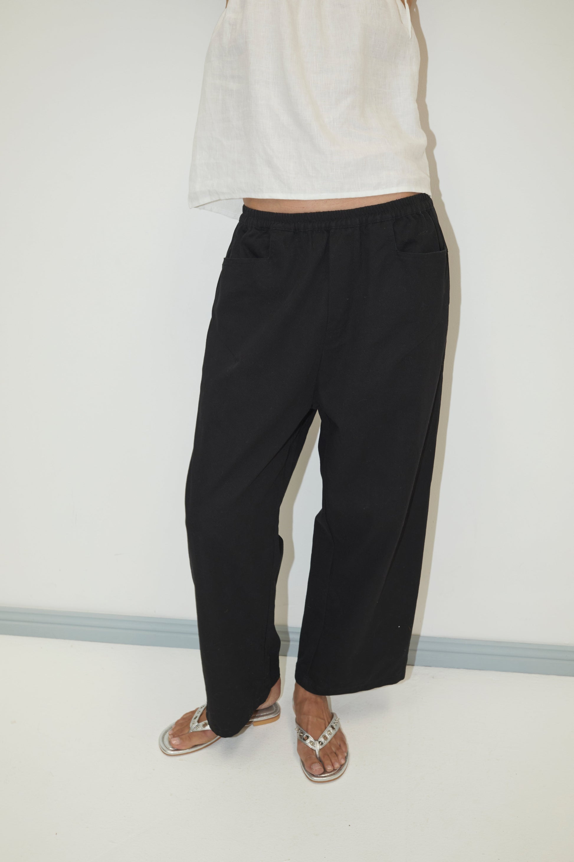 A person stands against a plain white wall, their head and most upper body out of frame, wearing a white sleeveless top, Square Pocket Pant - Black in relaxed-fit organic cotton twill, and metallic sandals.