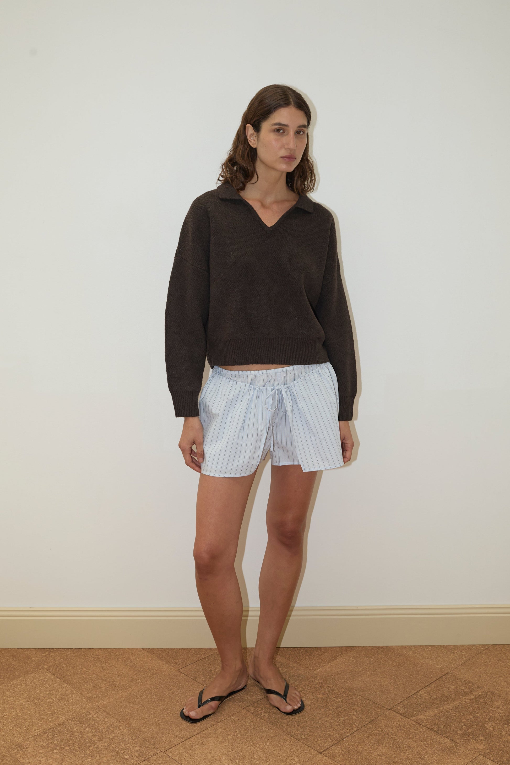 A woman stands indoors against a plain white wall, wearing the Deiji Studios Collared Pullover - Burnt Wood, light blue striped shorts, and black flip-flops. She has shoulder-length brown hair and a neutral expression.