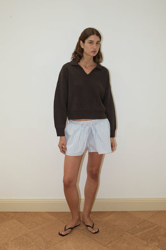 A woman stands indoors against a plain white wall, wearing the Deiji Studios Collared Pullover - Burnt Wood, light blue striped shorts, and black flip-flops. She has shoulder-length brown hair and a neutral expression.