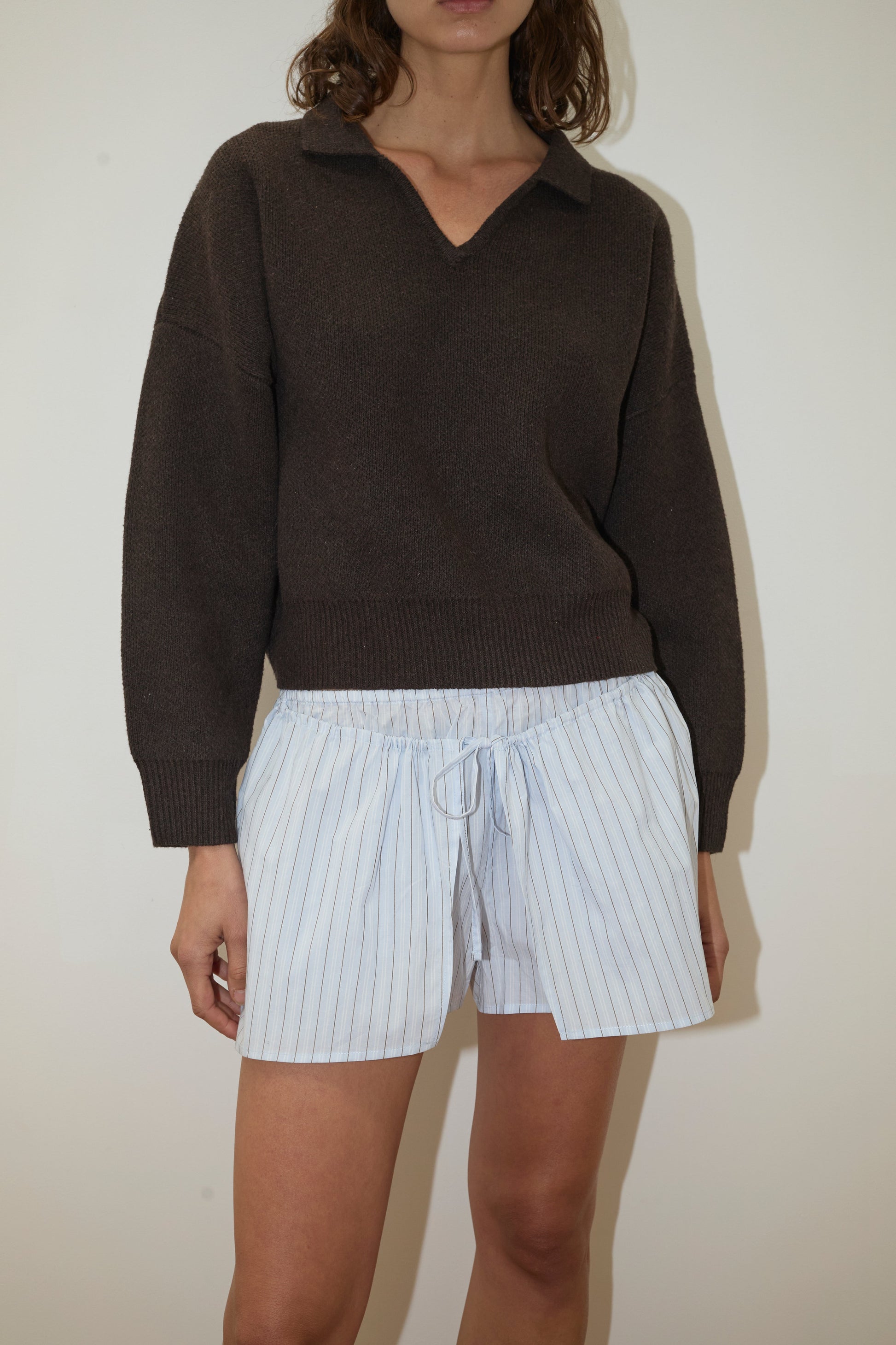 A woman wears the Collared Pullover - Burnt Wood by Deiji Studios, a dark brown relaxed long-sleeve knit, styled with white pinstriped shorts. She stands against a plain white background, her face partially out of frame.