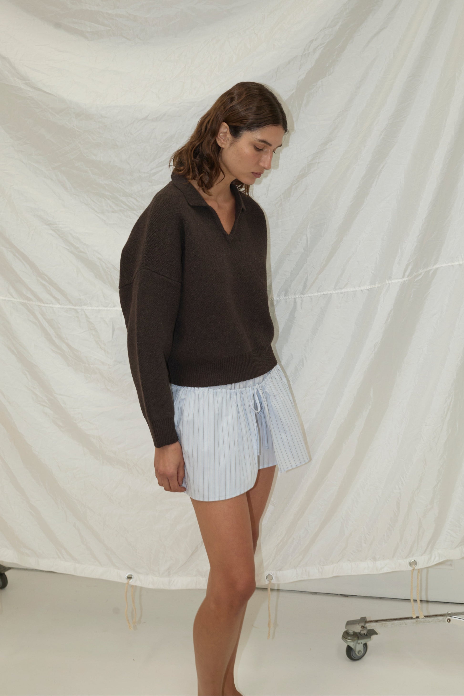 A woman with wavy brown hair wears the Collared Pullover - Burnt Wood by Deiji Studios and loose, light blue striped shorts, standing in front of a white wrinkled backdrop with metal stands visible at the bottom.