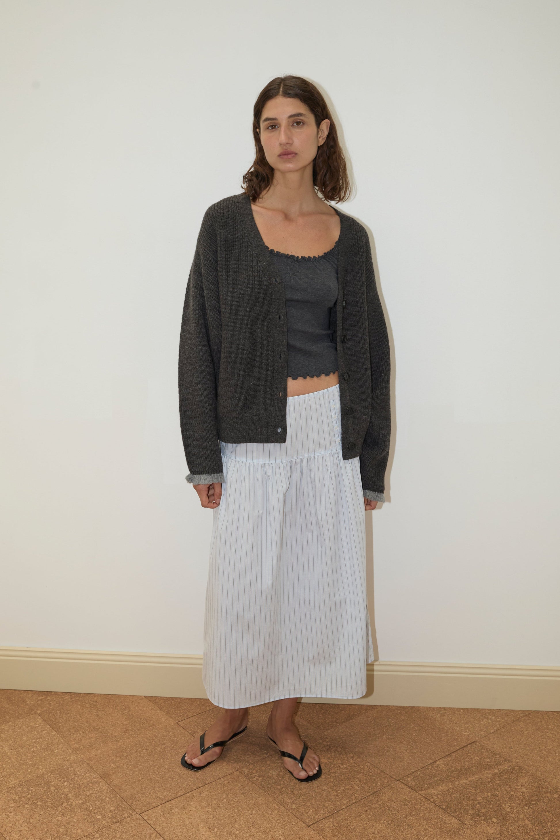 Indoors, a woman with shoulder-length brown hair and a neutral expression wears the Flounce Cardigan - Charcoal by Deiji Studios over a matching crop top, a long white striped skirt, and black flip-flops.