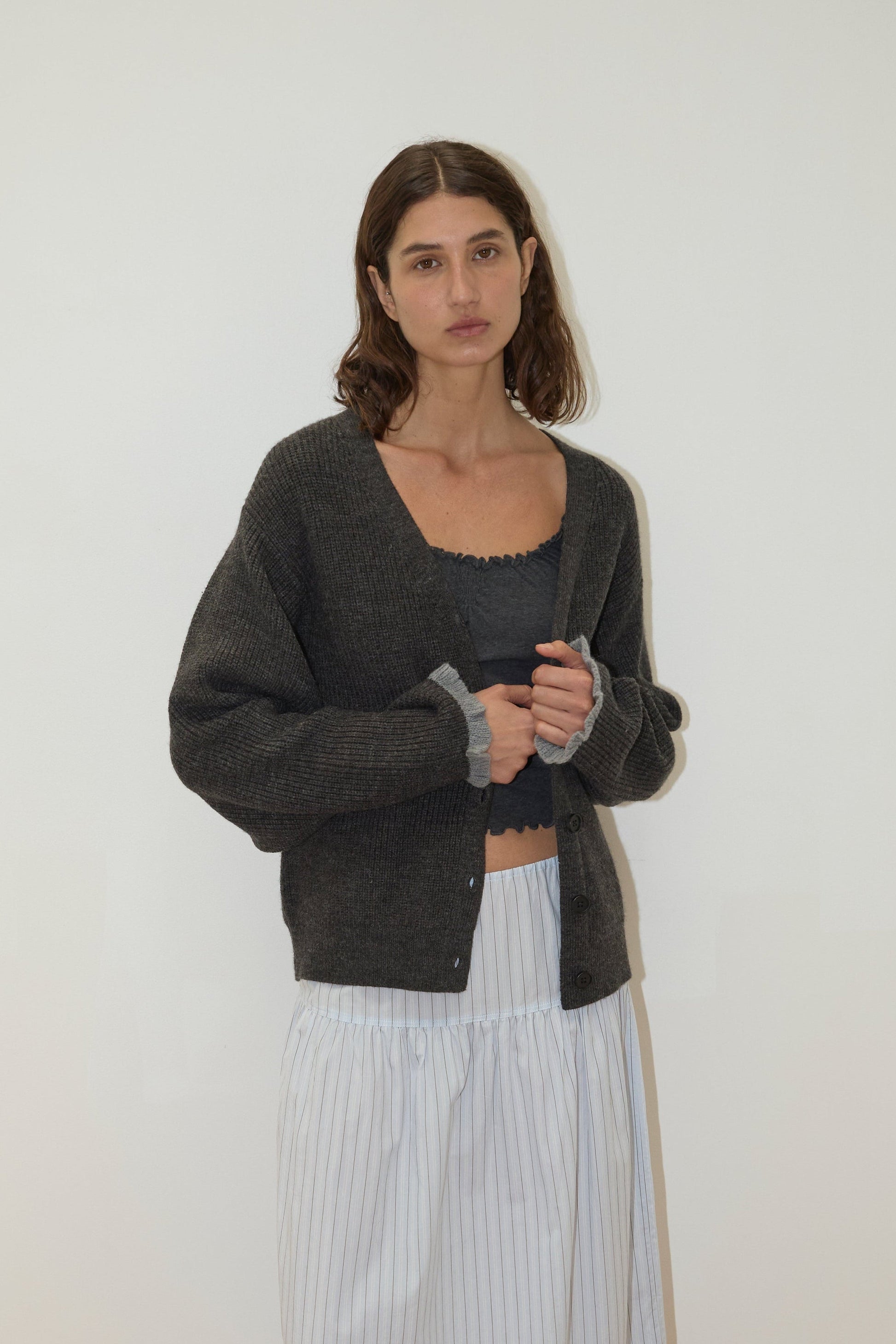 A woman with shoulder-length brown hair wears the Flounce Cardigan - Charcoal by Deiji Studios over a dark crop top and a long, white, lightly striped skirt. She stands against a plain light background, looking at the camera.
