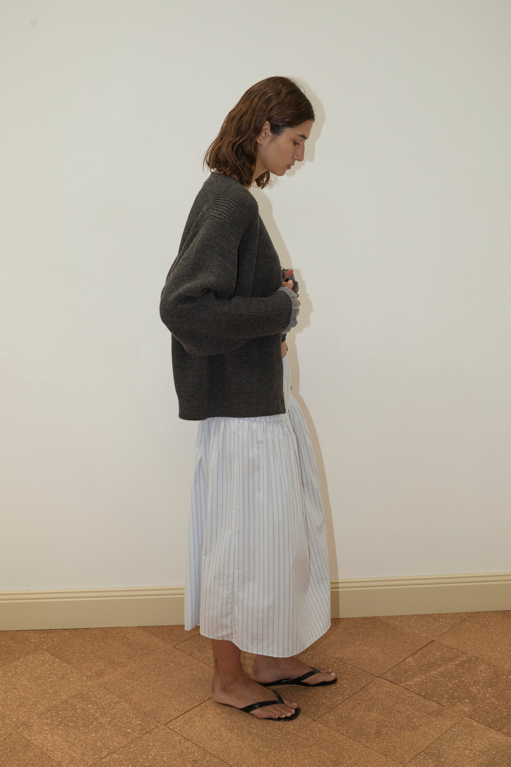 A person with shoulder-length brown hair stands sideways against a plain wall, wearing a Deiji Studios Flounce Cardigan - Charcoal, a long light blue striped skirt, and black sandals.