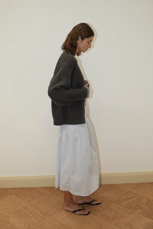 A person with shoulder-length brown hair stands sideways against a plain wall, wearing a Deiji Studios Flounce Cardigan - Charcoal, a long light blue striped skirt, and black sandals.