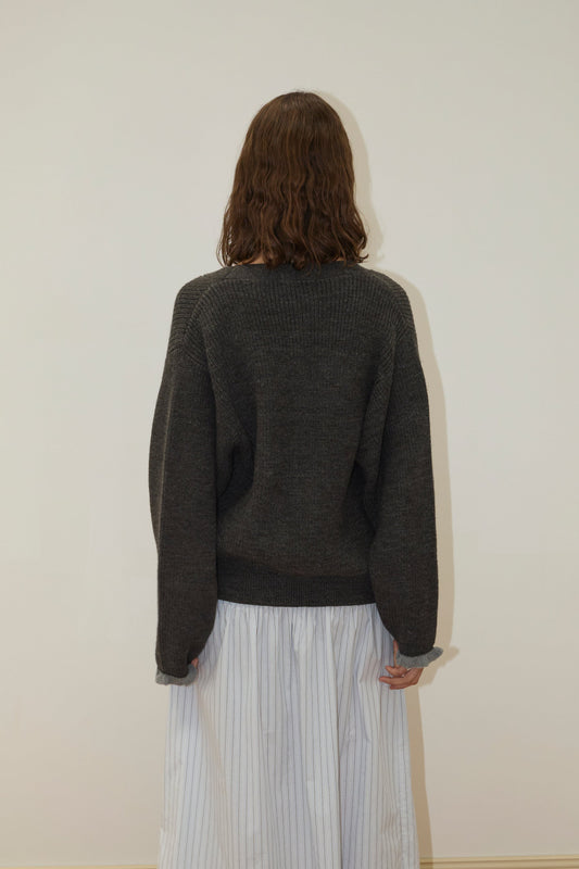 A person with shoulder-length brown hair faces away, wearing the Flounce Cardigan - Charcoal by Deiji Studios and a white skirt with thin vertical stripes, set against a plain light-colored wall.