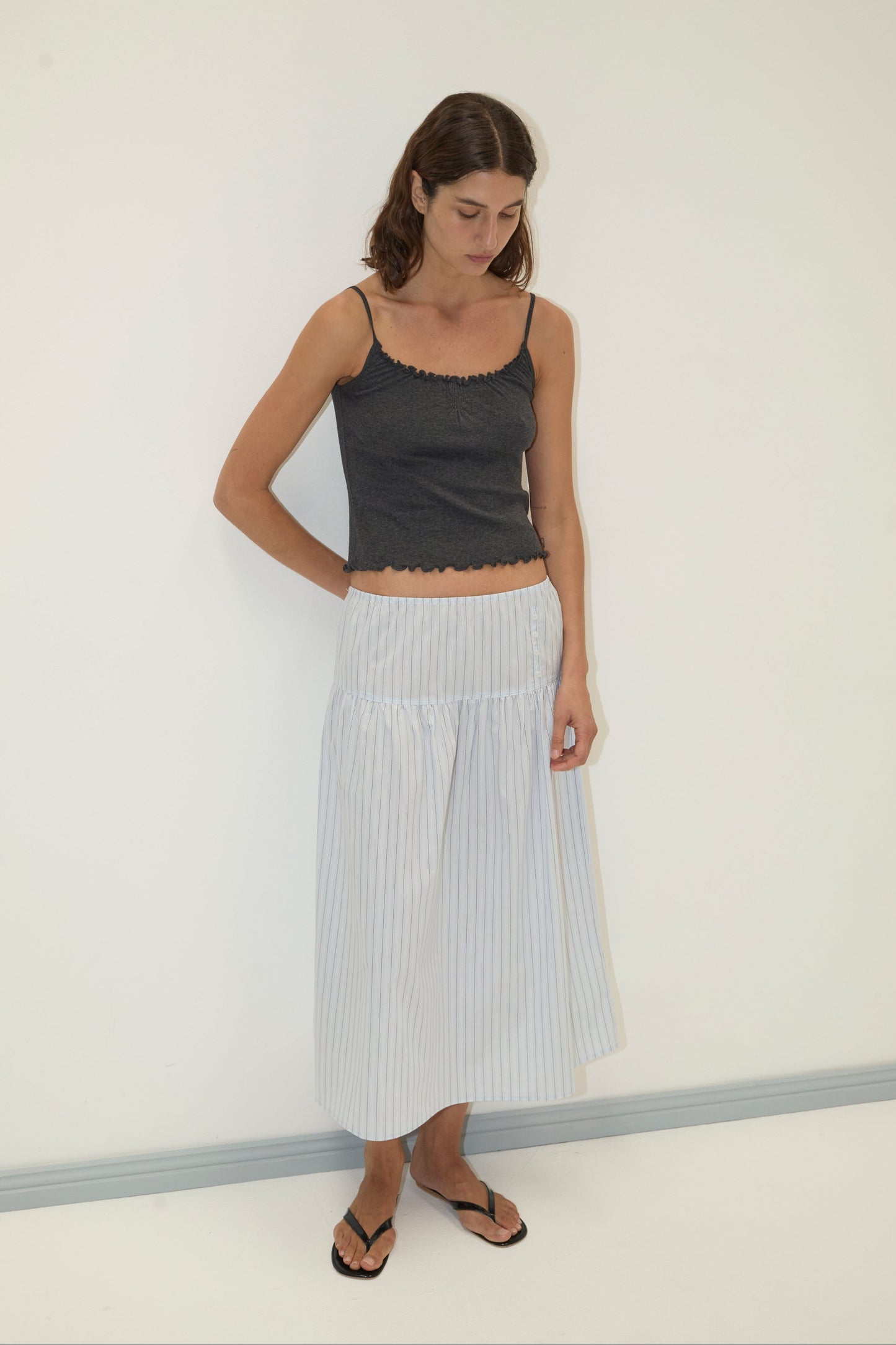 A woman stands against a plain white wall, wearing a dark gray sleeveless top, the Button Skirt—a light-colored striped midi skirt—and black flip-flops. She looks down with her arms behind her back.