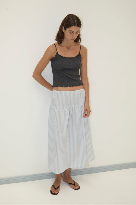 A woman stands against a plain white wall, wearing a dark gray sleeveless top, the light blue and white striped Button Skirt, and black flip-flops. She looks down with her hands behind her back.