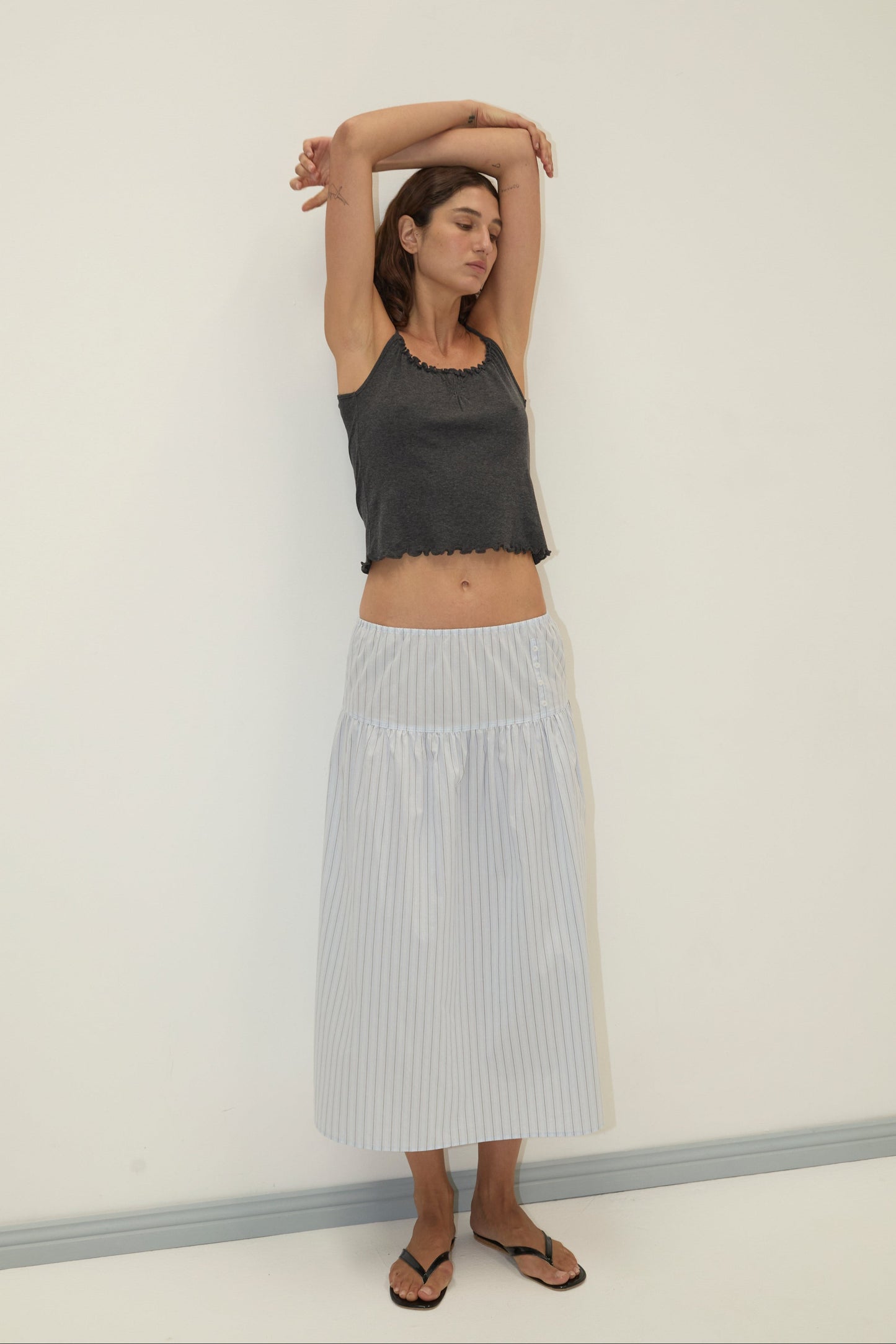 A woman stands against a plain white wall, wearing a dark sleeveless top and the Button Skirt - Mineral Stripe, a light blue and white striped mid-length skirt with an elasticated waist. She has black flip-flops on, arms raised and eyes closed, looking relaxed.