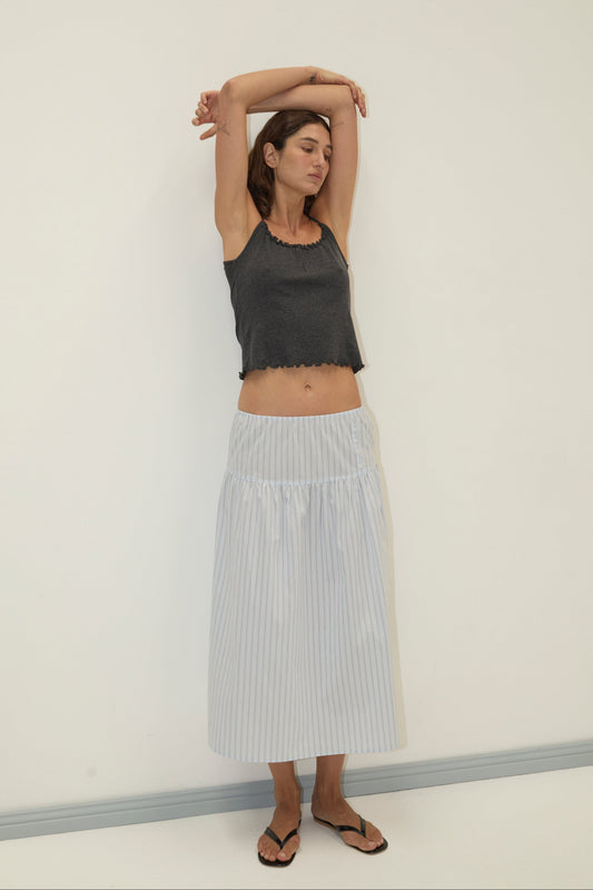 A woman stands against a plain white wall, wearing a dark sleeveless top and the Button Skirt - Mineral Stripe, a light blue and white striped mid-length skirt with an elasticated waist. She has black flip-flops on, arms raised and eyes closed, looking relaxed.