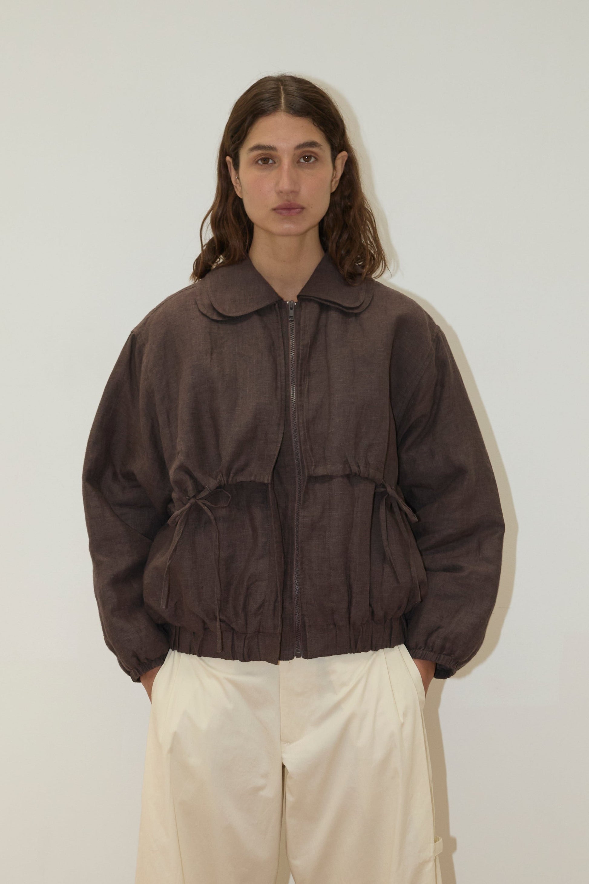 A person with wavy brown hair wears the Double Layered Jacket - Bark with a collar and light beige wide-leg pants, standing against a plain off-white background.