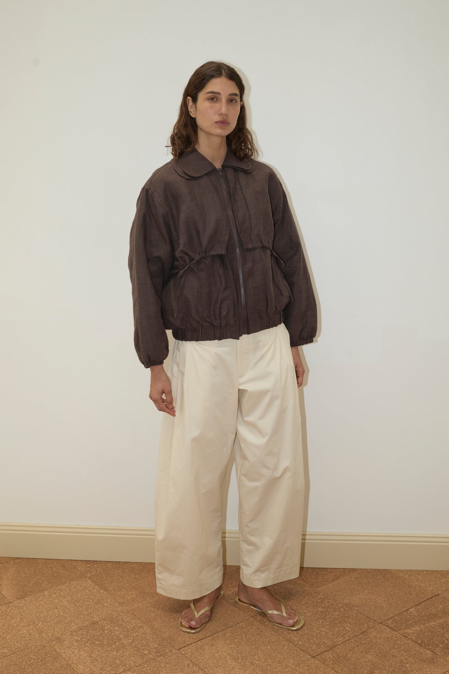 A person with wavy brown hair wears the Double Layered Jacket - Bark, wide cream pants, and tan sandals, standing against a plain white wall and brown tiled floor.