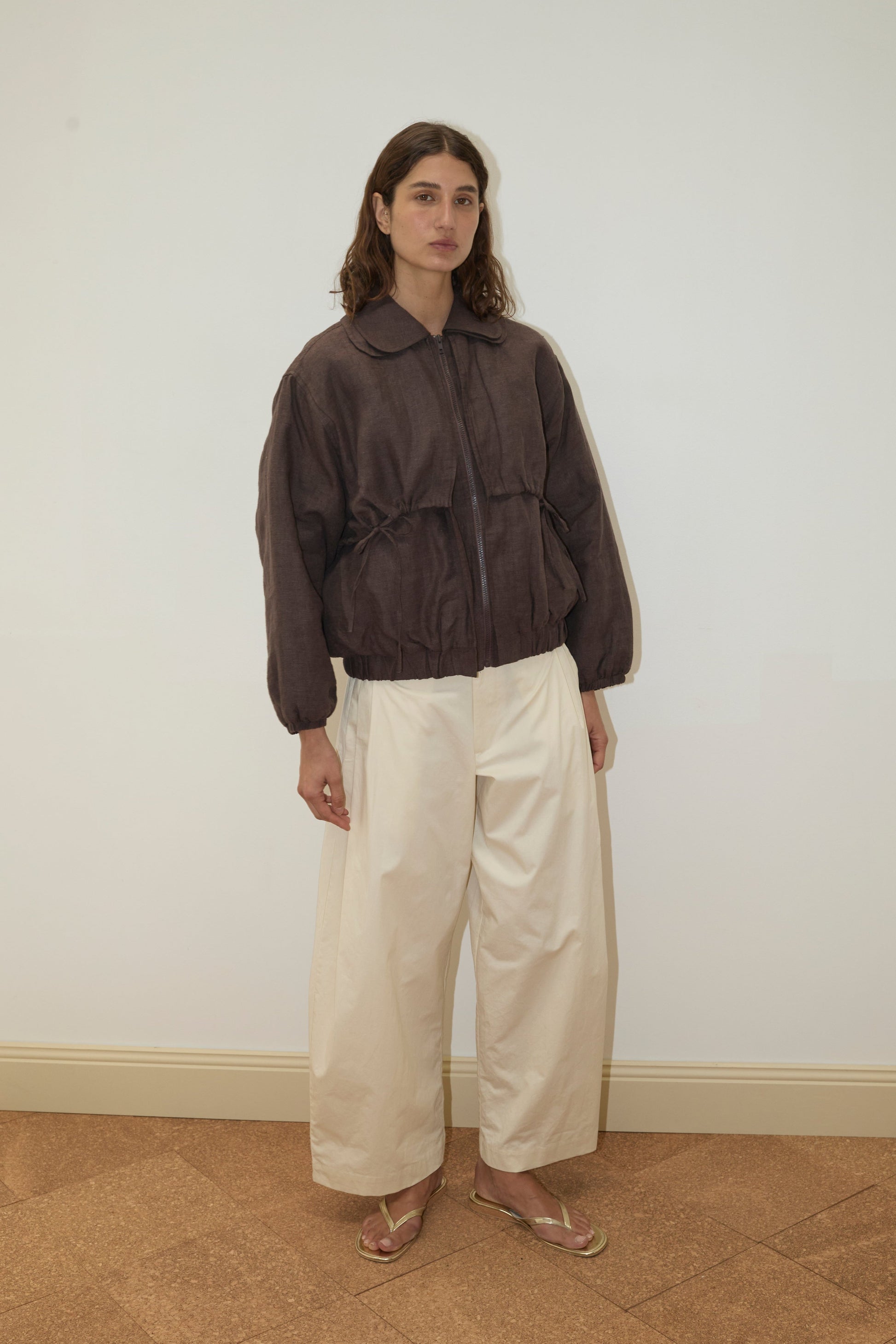 A person with wavy brown hair wears the Double Layered Jacket - Bark, wide cream pants, and tan sandals, standing against a plain white wall and brown tiled floor.