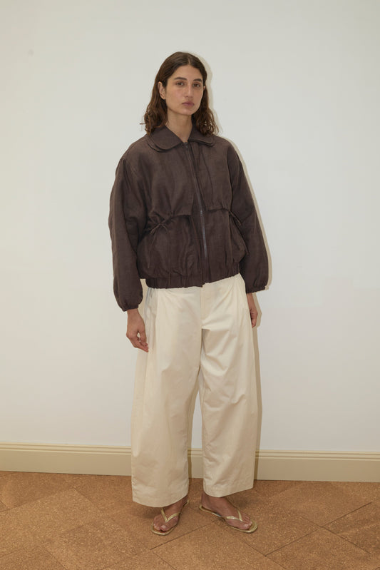 A person with wavy brown hair wears the Double Layered Jacket - Bark, wide cream pants, and tan sandals, standing against a plain white wall and brown tiled floor.