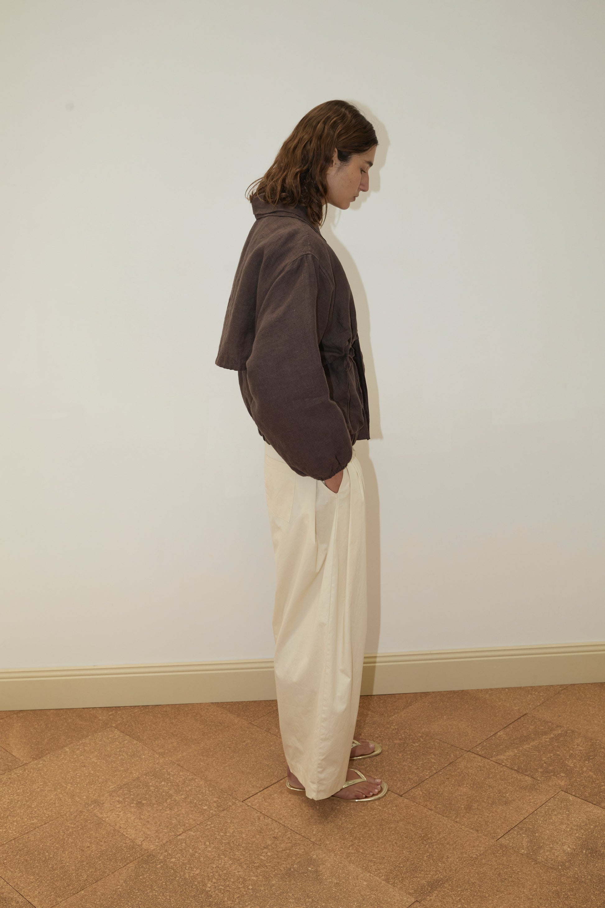 A person with shoulder-length brown hair stands sideways against a plain wall, wearing the Double Layered Jacket - Bark, cream-colored pants, and sandals on a patterned brown floor.