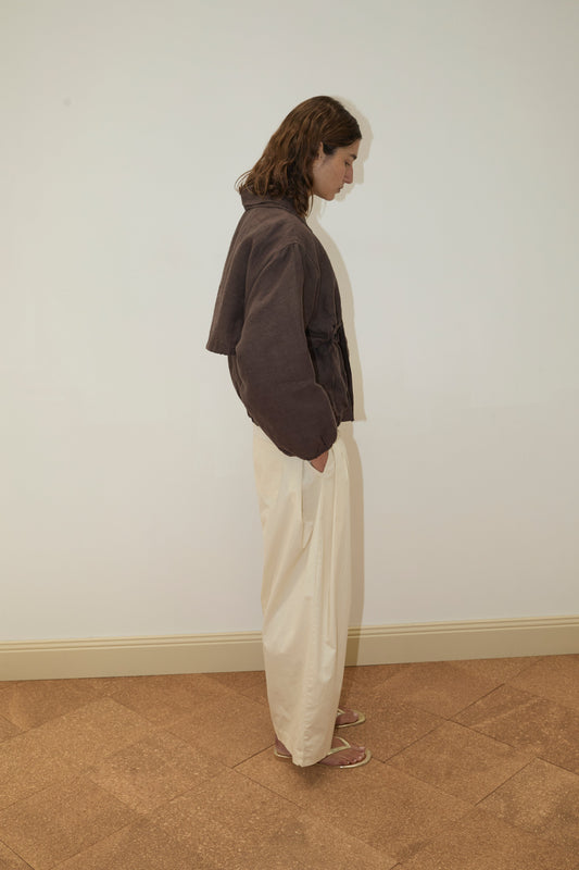 A person with shoulder-length brown hair stands sideways against a plain wall, wearing the Double Layered Jacket - Bark, cream-colored pants, and sandals on a patterned brown floor.