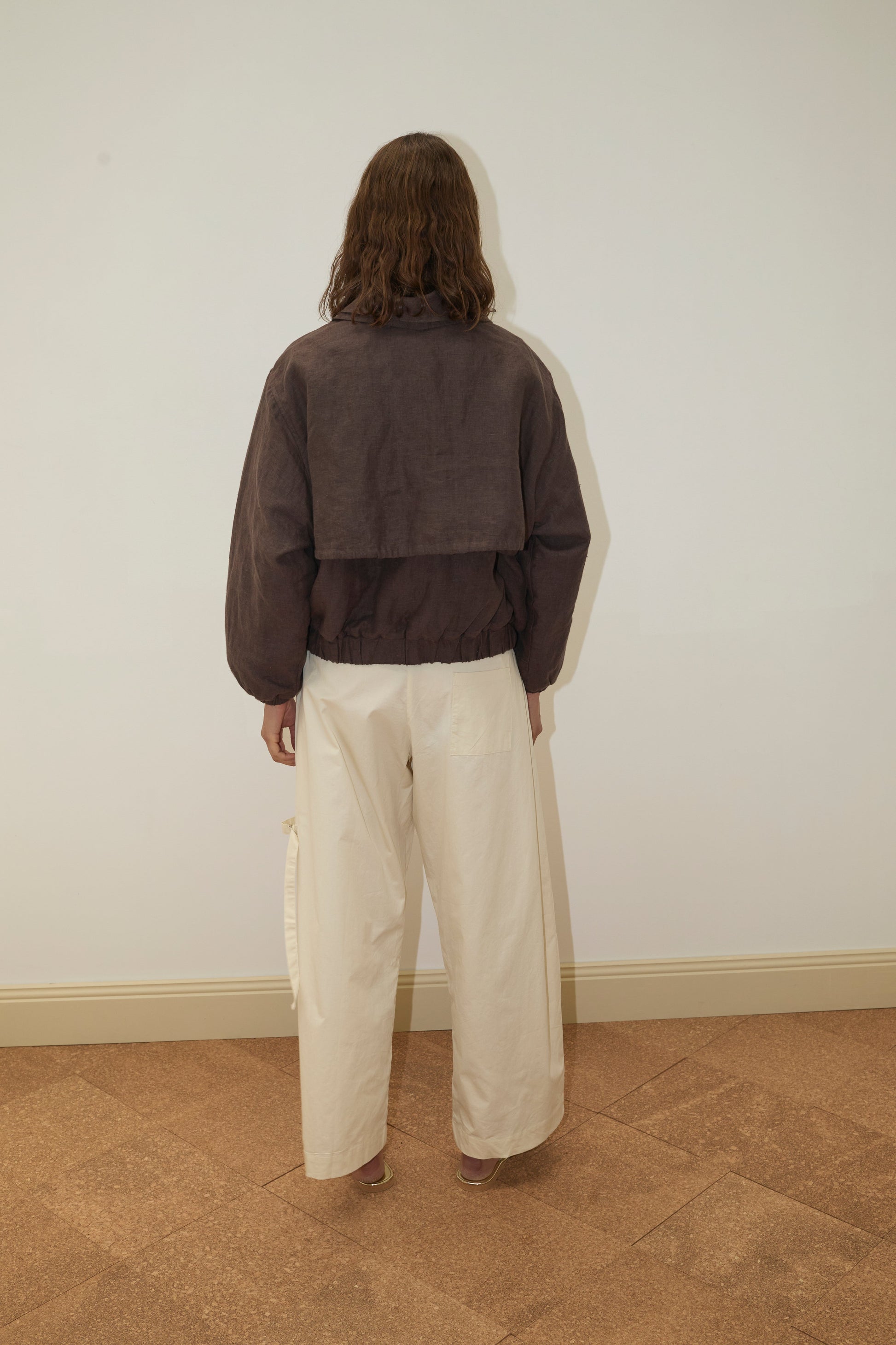 A person with shoulder-length brown hair stands facing a plain white wall, wearing the Double Layered Jacket - Bark and wide-leg cream-colored pants on a light brown tiled floor.