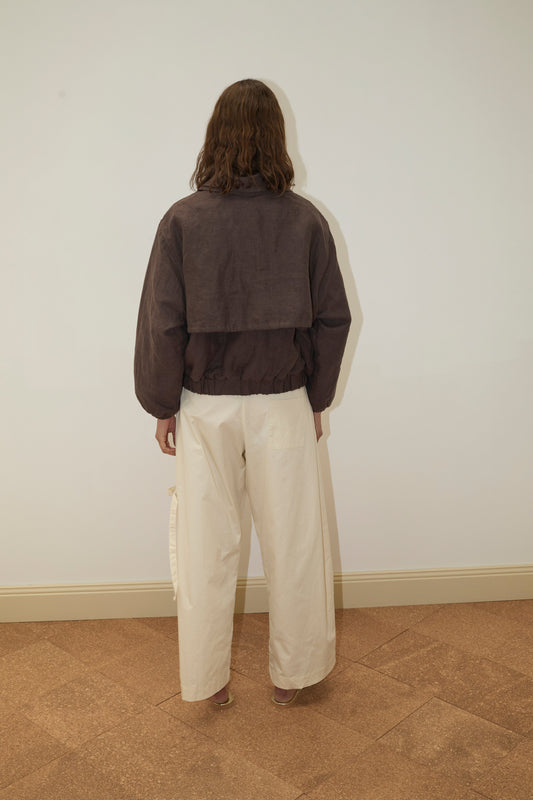 A person with shoulder-length brown hair stands facing a plain white wall, wearing the Double Layered Jacket - Bark and wide-leg cream-colored pants on a light brown tiled floor.