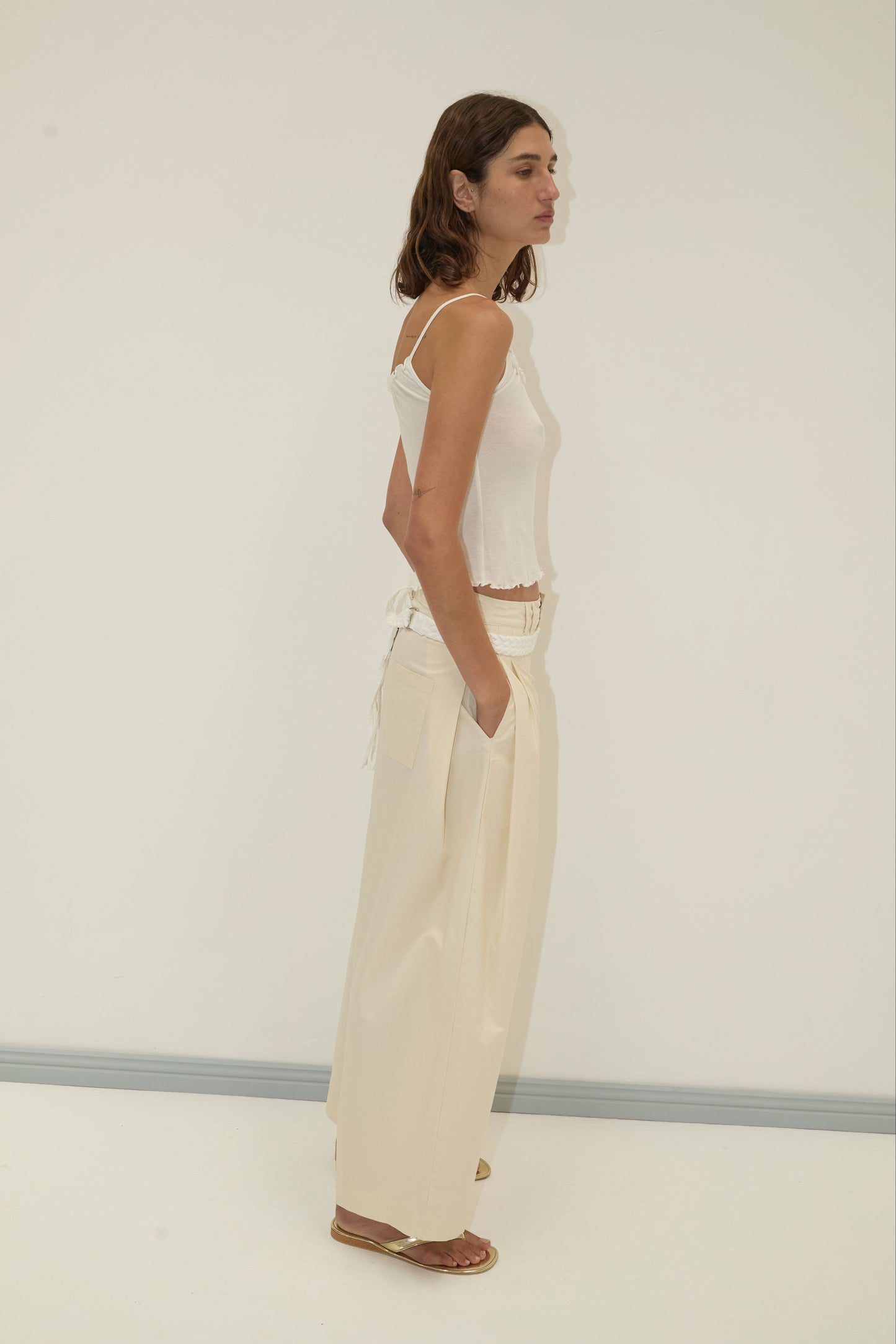 A woman with shoulder-length brown hair stands sideways against a light wall, wearing a white tank top and Deiji Studios Tab Pant - Off White with hands in pockets and brown sandals, looking relaxed.