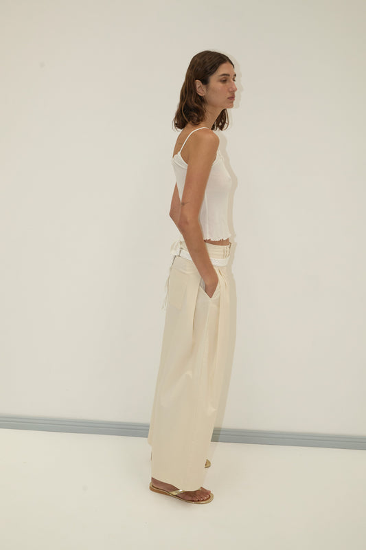 A woman with shoulder-length brown hair stands sideways against a light wall, wearing a white tank top and Deiji Studios Tab Pant - Off White with hands in pockets and brown sandals, looking relaxed.