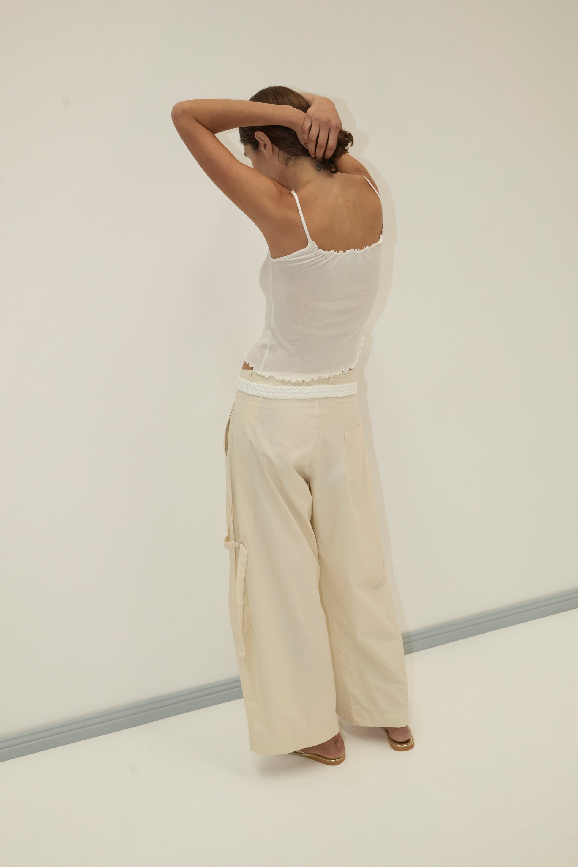 A person stands with their back to the camera, arms raised and hands behind their head. They wear a white tank top and the Tab Pant - Off White by Deiji Studios in a minimal, light-colored room.
